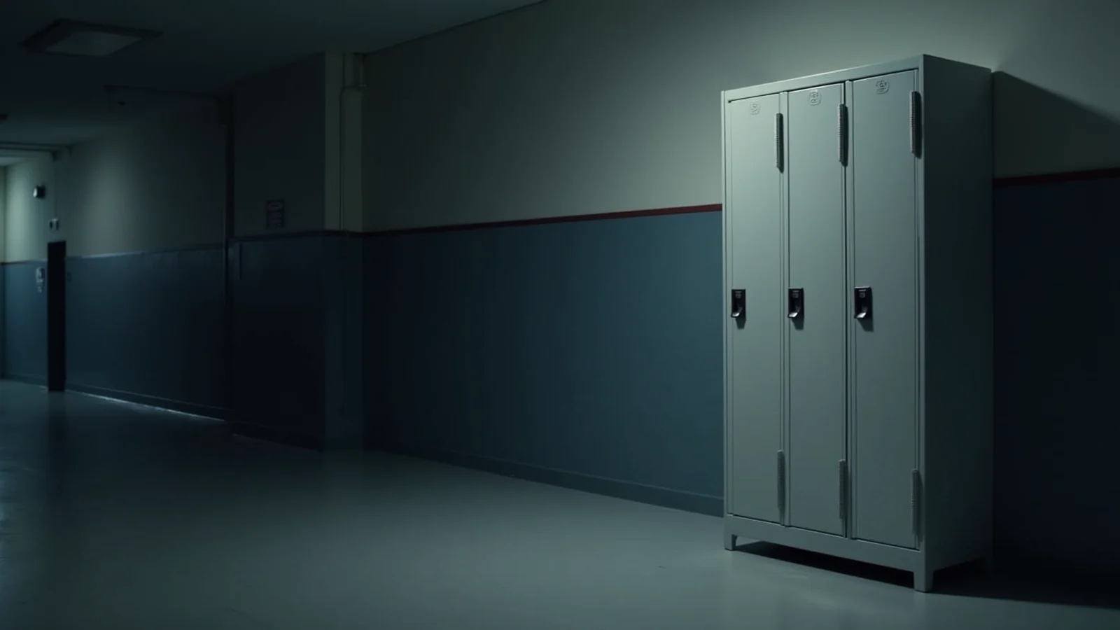 A bank of pale grey steel school lockers standing against the wall of a dim empty corridor, lit cold from one side.