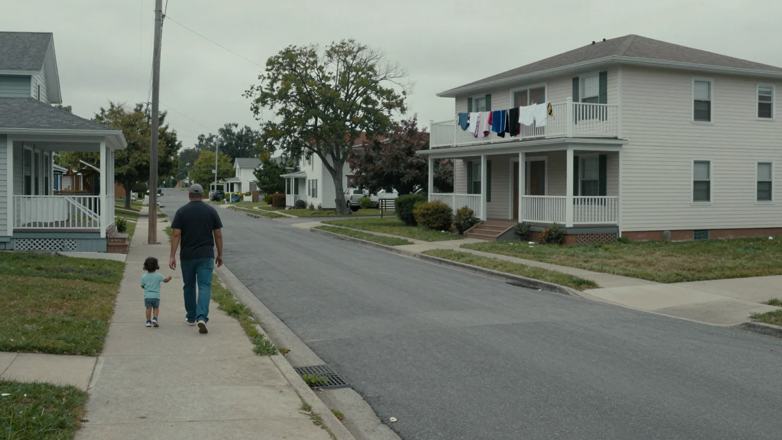 A parent and a small child walking away along the sidewalk of a quiet street of modest houses, laundry drying on a balcony rail, under an overcast sky.