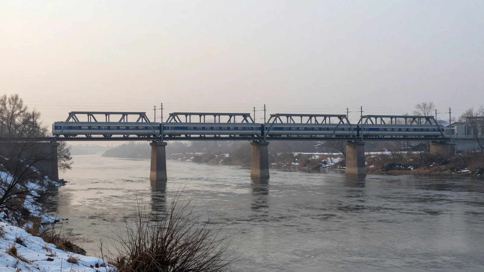 A silver passenger train crossing a steel truss bridge over a misty river at dawn, with snow along the banks.