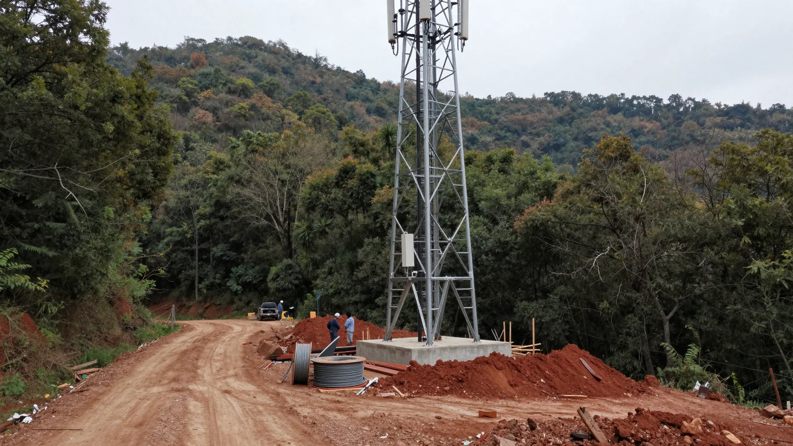 A galvanised steel lattice tower stands on a fresh concrete pad cut into a wooded hillside, photographed from a red dirt access track under a flat grey sky. Mounded red earth, a large spool of heavy cable and stacked timber lie at its base; two small figures in hard hats stand beside a pickup truck further down the track.