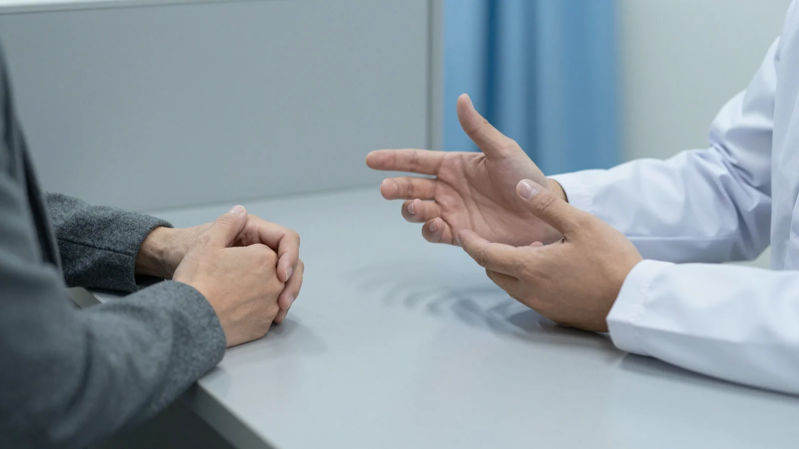Cropped view of clasped hands and a white-coated person's gesturing hands across a grey consultation table.