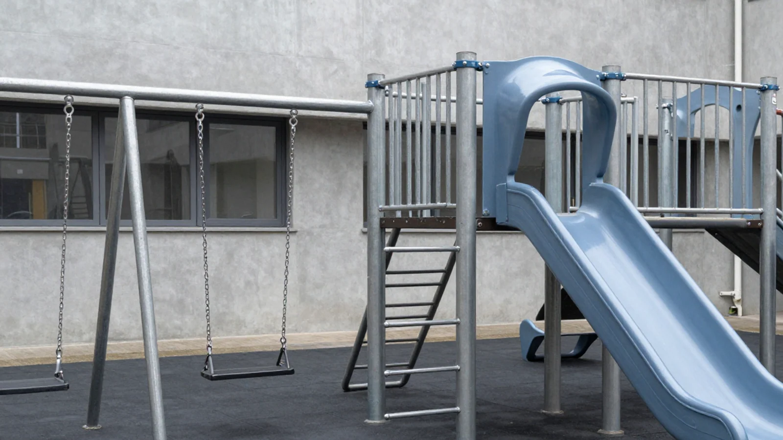 Empty swings, a ladder and a blue slide stand beside a grey building.