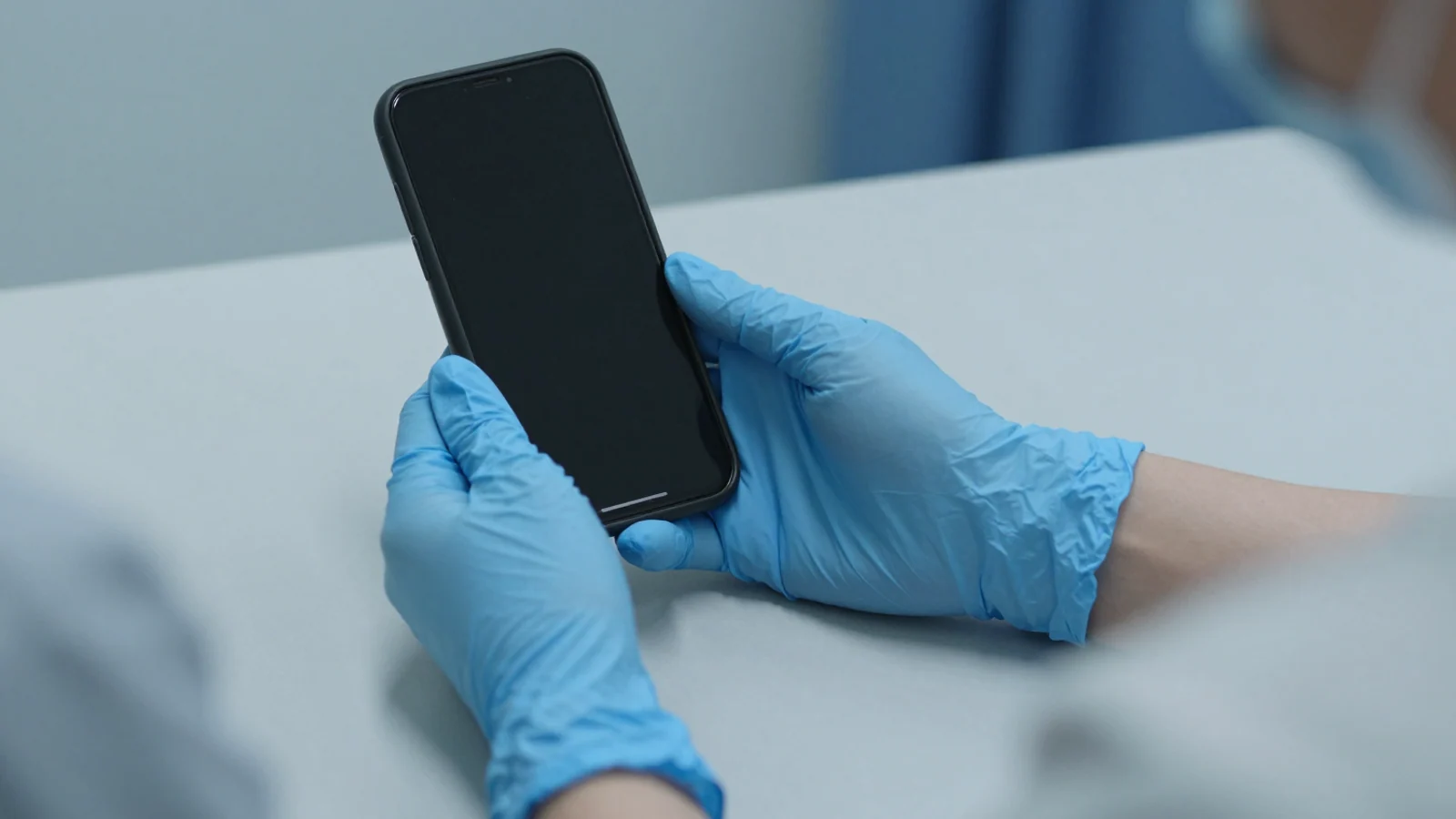 Blue-gloved hands hold a black phone with an unlit screen above a pale consultation table.