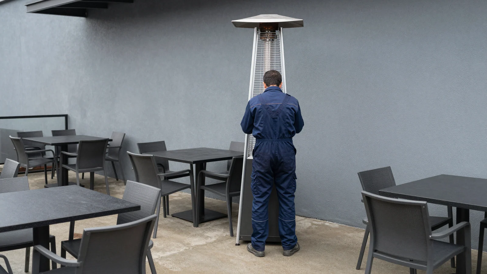 A worker in blue coveralls faces a tall patio heater among empty grey tables and chairs on a covered terrace.