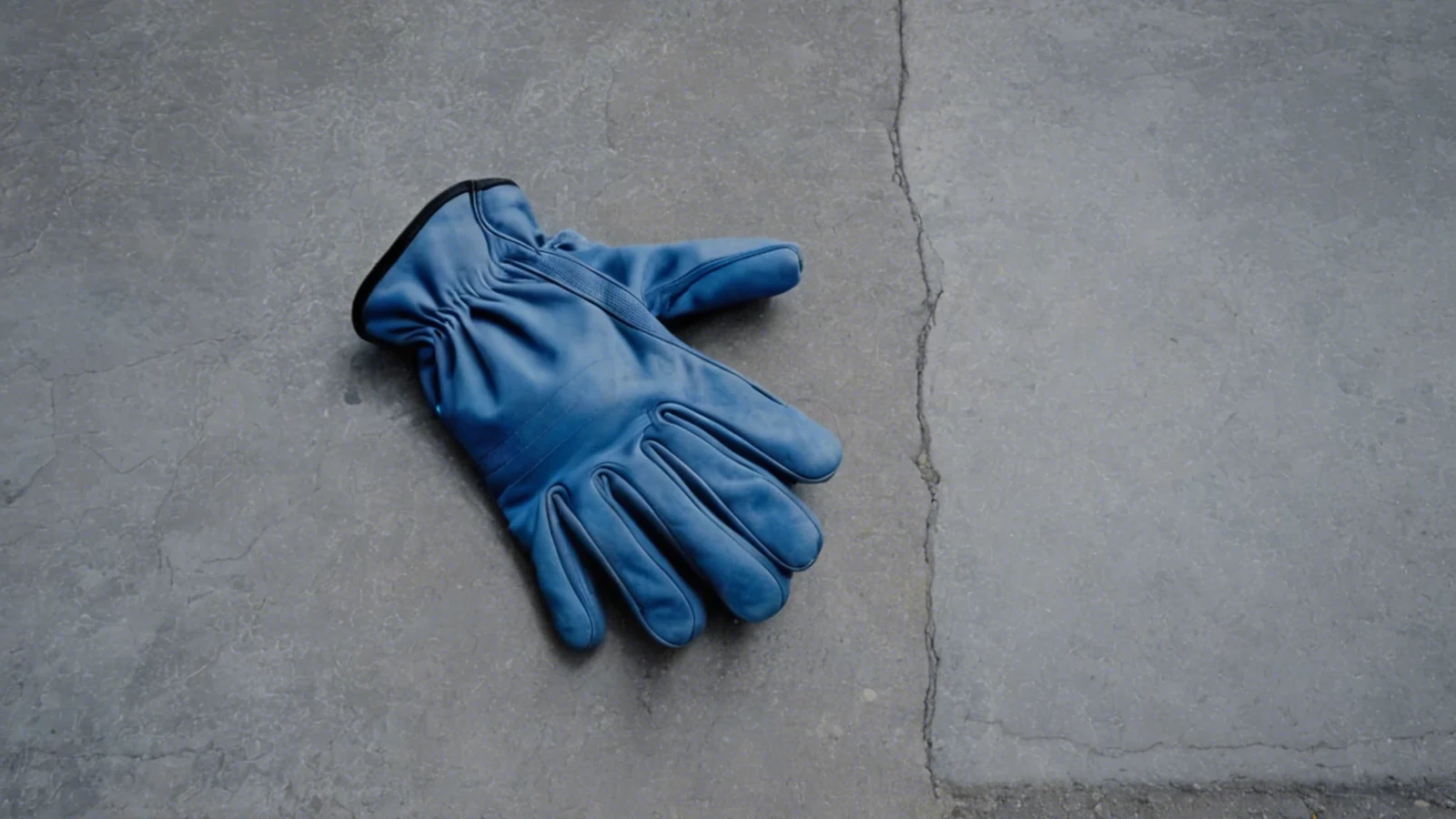 A pair of worn blue work gloves lying palm-down and empty on a cracked grey concrete slab in cold flat light, with nothing else in the frame.