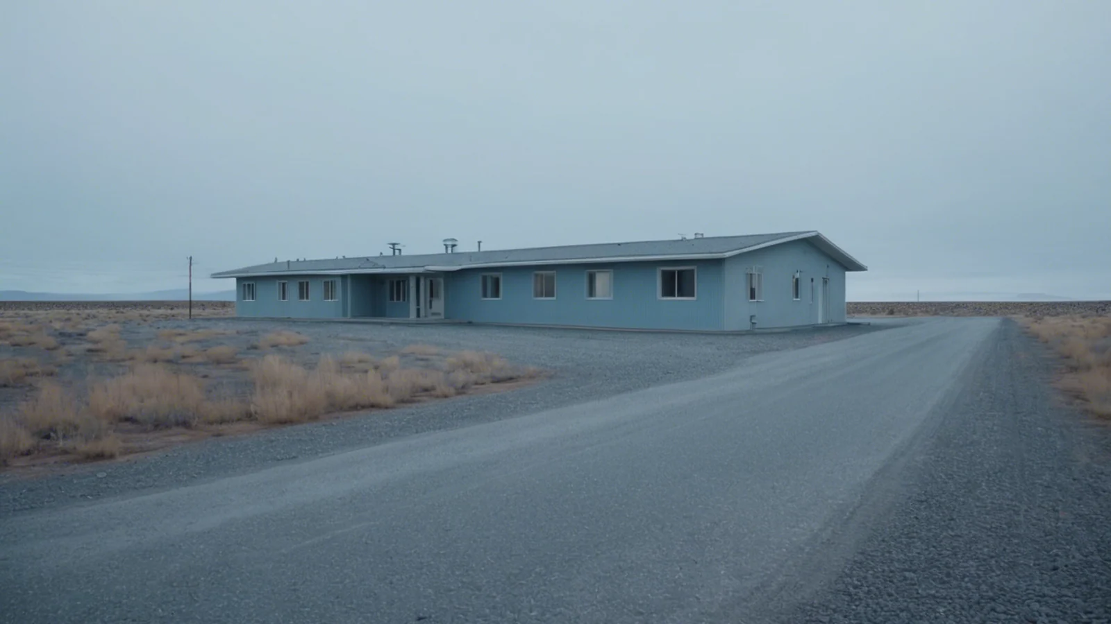 A long empty grey road running straight across flat open scrubland under a low overcast sky toward a single low building standing alone on the horizon, with no sign, vehicle or person anywhere in the frame.