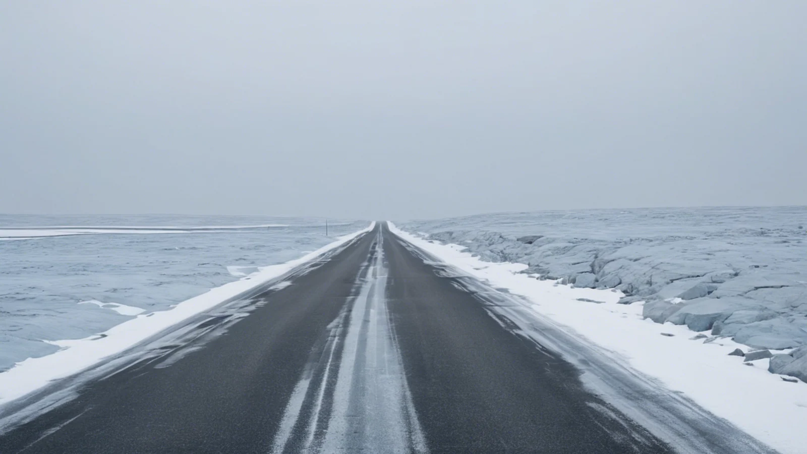 A straight dark road recedes across a flat snow-covered plain beneath a pale overcast sky.