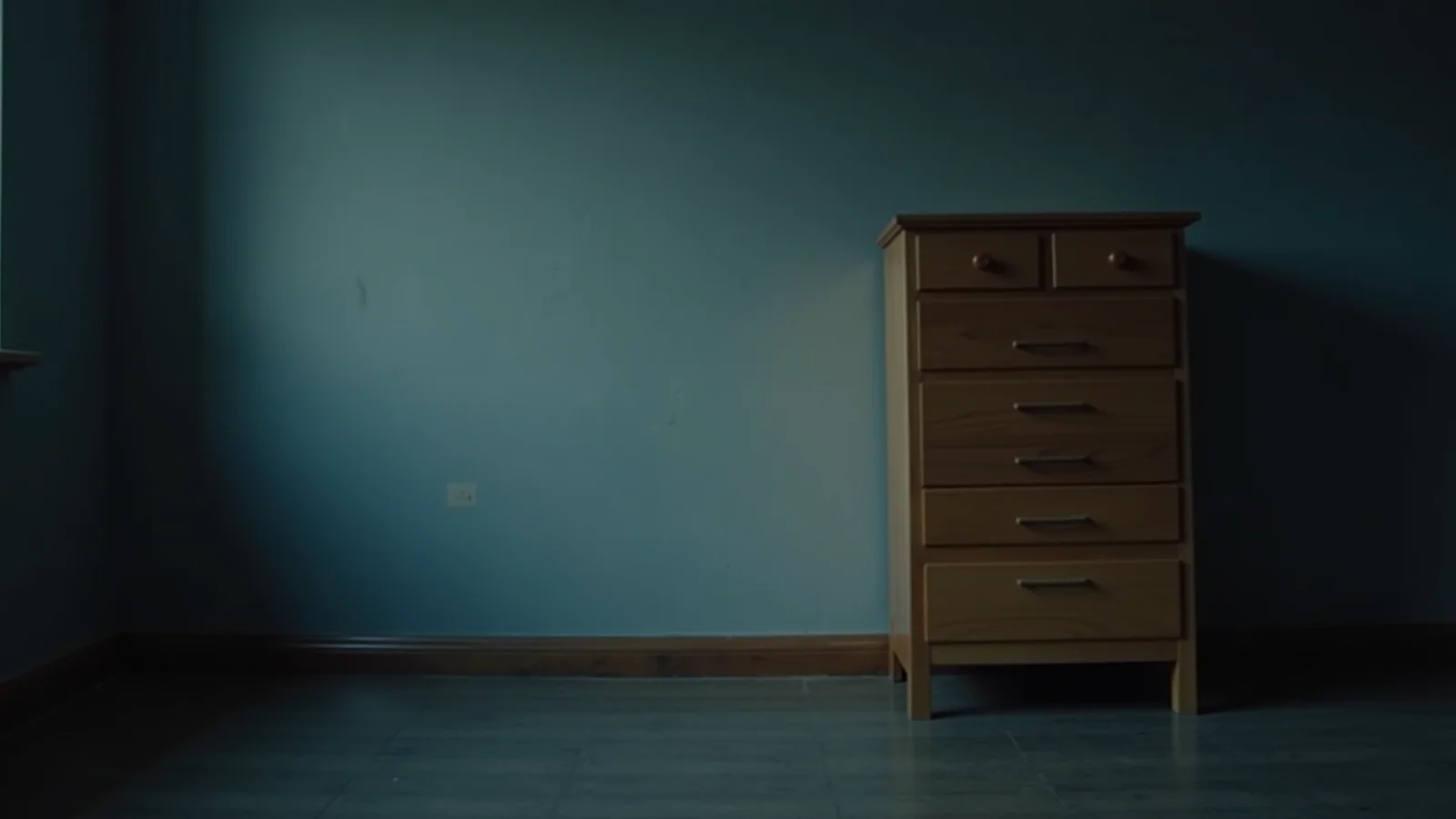 A plain wooden chest of drawers standing alone against a bare wall in a dim room.